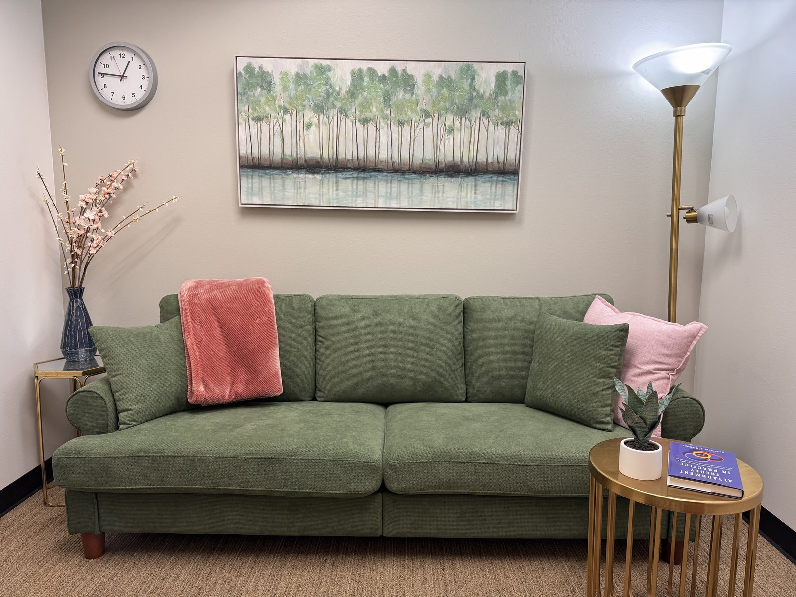 A calm therapy office: a sage-green sofa with blush and pink pillows, warm lamplight, cherry blossoms, and a painting of trees reflected in water.